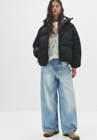 Crna puffer jakna s kapuljačom, siva majica s grafikom, preveliki svijetloplavi traperice i smeđe čizme s potpeticama. Glatke teksture, opušten kroj.