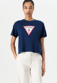 Tamsiai mėlynas trumpintas marškinėliai, pagaminti iš švelnios medžiagos, su raudonu ir baltu trikampiu logotipu, kuriame užrašyta "GUESS" ir klaustukas.