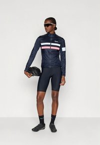 Temno modra kolesarska jakna z roza in belimi progami, iz lahkega materiala, v kombinaciji s črnimi kratkimi hlačami, nogavicami in čevlji, drži kolesarsko čelado.