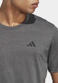 Camiseta de algodón gris con cuello redondo y un logo de tres franjas negras en el área del pecho izquierdo. Mangas cortas con una textura suave.