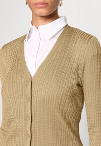 Beige pletený kardigan s texturovaným žebrovaným vzorem, designem s V výstřihem a zapínáním na knoflíky, nošený přes bílou košilí s límečkem.