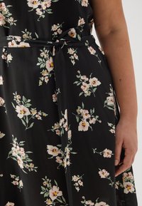 Robe noire à motifs floraux avec des fleurs roses et jaunes, dotée d'une ceinture nouée à la taille et d'une texture de tissu lisse.