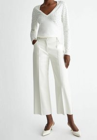 White long-sleeve sweater with V-neck and silver studs, paired with wide-leg white trousers. Features a button closure and side pockets.