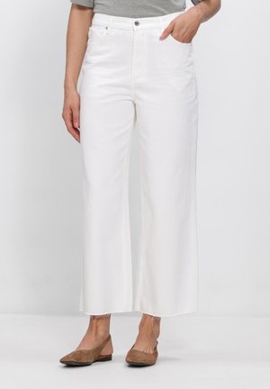 Vrouw draagt hoge taille witte jeans met wijde pijpen en onafgewerkte zoom, grijze top en bruine platte slingback schoenen, staand tegen een witte achtergrond.