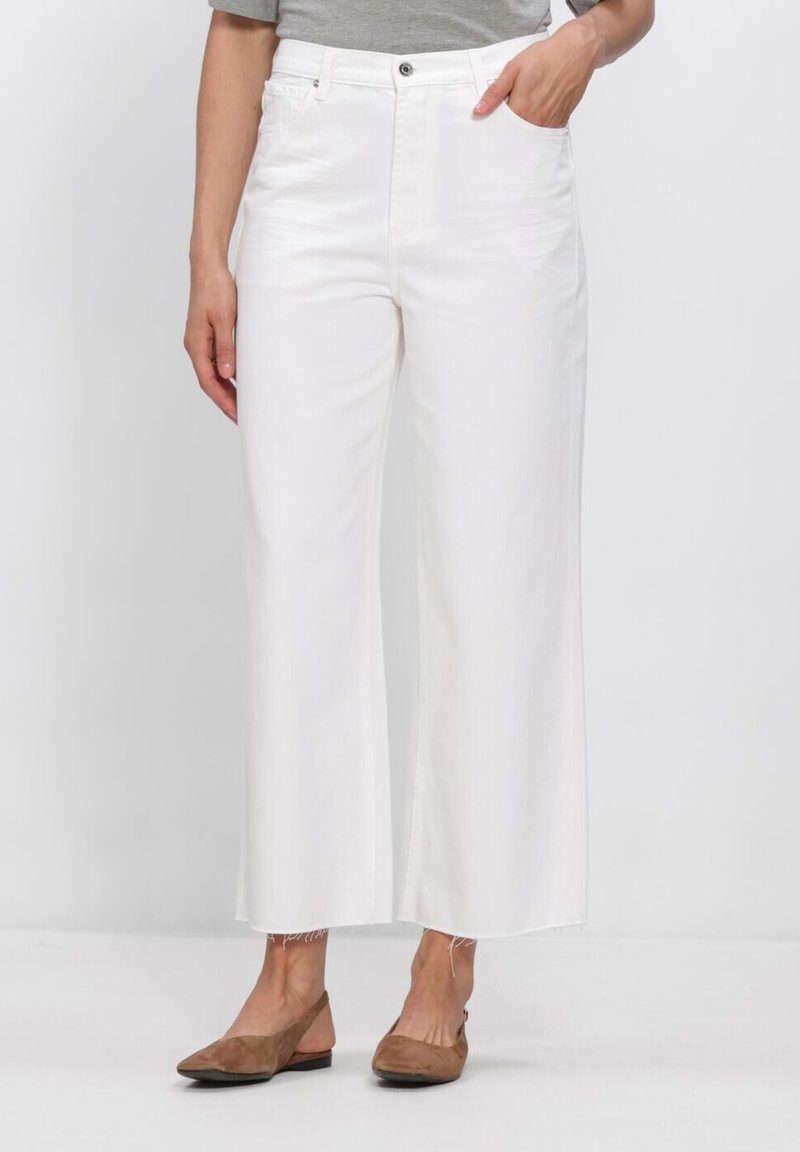 Woman wearing high-waist white wide-leg jeans with raw hem, gray top, and brown flat slingback shoes, standing against a white background.
