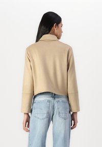 Veste beige courte en matière lisse, avec un col cranté et des manches larges. Portée par-dessus un jean bleu clair, ample.