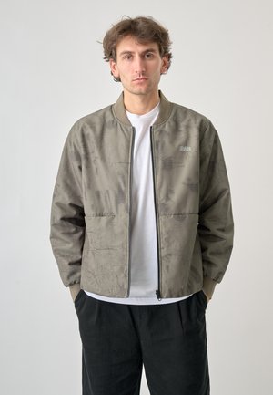 Olivgrüner Bomberjacke mit strukturiertem Muster, geripptem Kragen und einem Frontreißverschluss. Kombiniert mit einem weißen T-Shirt und schwarzen Hosen.