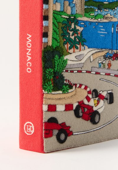 Κόκκινη ράχη βιβλίου με το κείμενο "MONACO", κάλυμμα από τσόχα που δείχνει έναν αγώνα της Φόρμουλα 1 με κόκκινα αυτοκίνητα, φοίνικες, θεατές και ένα παραθαλάσσιο αστικό τοπίο στο φόντο.