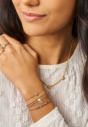 Frau trägt eine weiße, strukturierte Bluse, eine goldene Blumenkette, einen goldenen Blumenring und mehrere goldene Armbänder mit Herz- und Blumenanhängern.