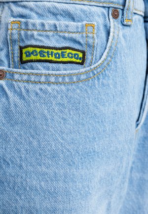 Helle blaue Jeans aus Denim mit einer Gesäßtasche, die mit einem bunten gestickten Etikett verziert ist. Gelbe Nähte und ein Standardknopfverschluss sind sichtbar.