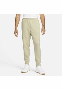 Světle béžové joggingové kalhoty vyrobené z měkké tkaniny, s elastickým pasem a manžetami na kotnících. Logo Nike je viditelné na horní levé straně.