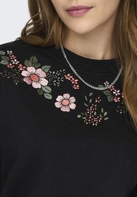 Sudadera negra con bordado floral en rosa y verde en el cuello, con un collar de cadena plateada visible sobre la tela.