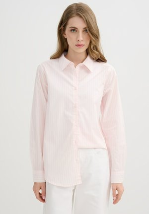 Mujer con cabello castaño claro que lleva una camisa rosa claro de manga larga con botones y pantalones blancos, de pie frente a un fondo blanco liso.