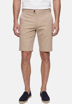 Homme portant un short beige au genou, une chemise blanche et des chaussures slip-on bleu marine, debout devant un fond clair uni.