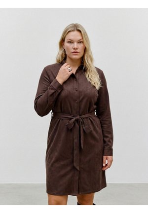 Femme aux longs cheveux blonds portant une robe marron boutonnée, nouée à la taille, debout devant un fond clair uni.