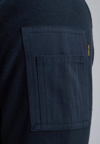 Marineblauw stoffen overhemd met een textuur gebreide body en een platte opgestikte zak op de schouder met lichte stiksels en een klein merklabel.