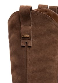 Brūni zamšādas zābaki ar gludu tekstūru, noapaļotu formu un cilpu akcentu. Iekļauj nelielu zīmola metāla detaļu tuvu augšdaļai.