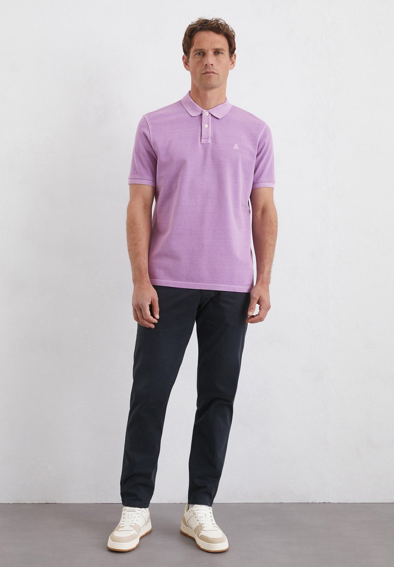 Koszulka polo z krótkim rękawem w jasnofioletowym kolorze, z wąskim kołnierzykiem, zapięciem na trzy guziki i małym logo. W zestawie z ciemnymi spodniami i białymi trampkami.