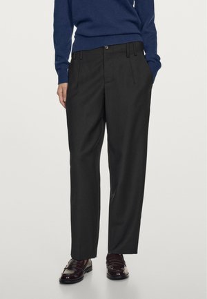 Personne portant un pantalon noir plissé, des mocassins en cuir marron foncé et un pull bleu à manches longues, avec une main dans la poche.