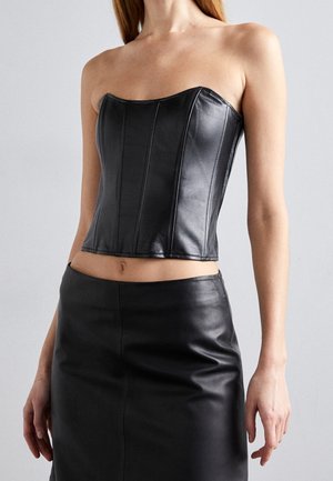 Femme portant un corset en cuir noir sans bretelles et une jupe en cuir noir assortie sur un fond gris uni.
