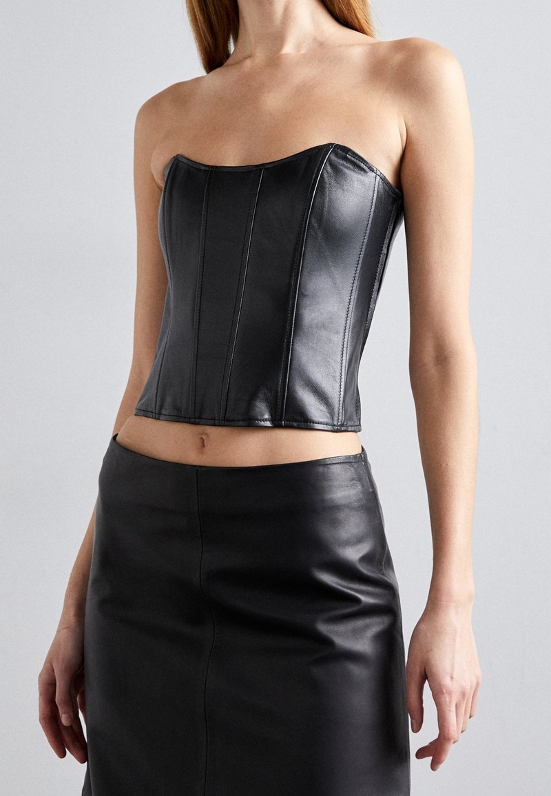 Femme portant un corset en cuir noir sans bretelles et une jupe en cuir noir assortie sur un fond gris uni.