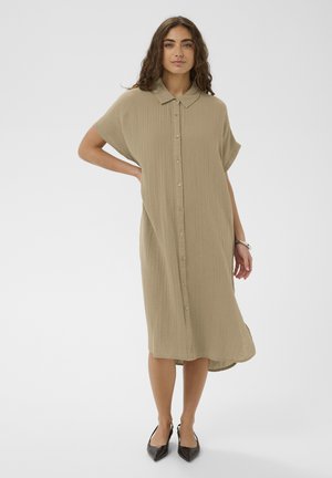 Donna con capelli lunghi e ricci che indossa un vestito midi beige morbido con bottoni e maniche corte, e scarpe nere con punte a punta, in piedi con una mano sui fianchi.