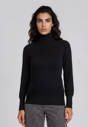 Vrouw met lang donker haar, gekleed in een strakke zwarte coltrui en grijze broek, staand tegen een egale lichtgrijze achtergrond.