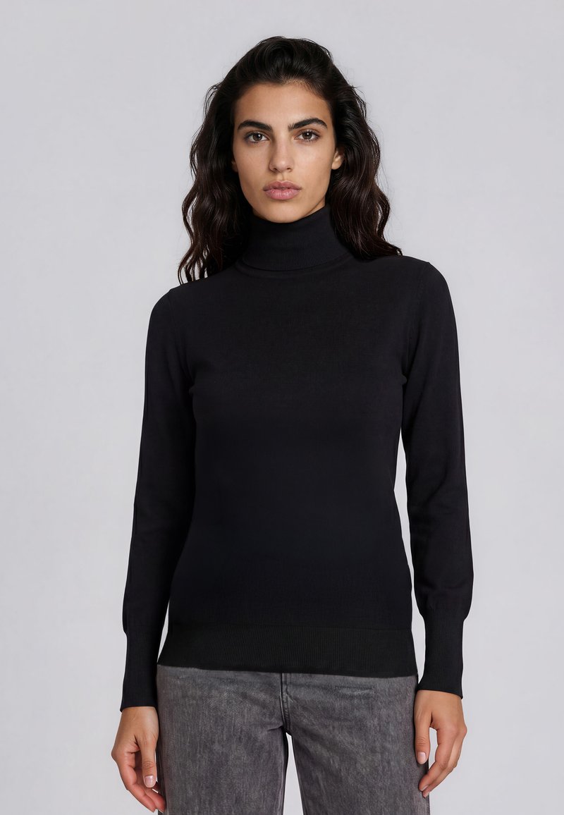 Femme aux longs cheveux foncés portant un pull noir ajusté à col roulé et un pantalon gris, debout devant un fond uni gris clair.