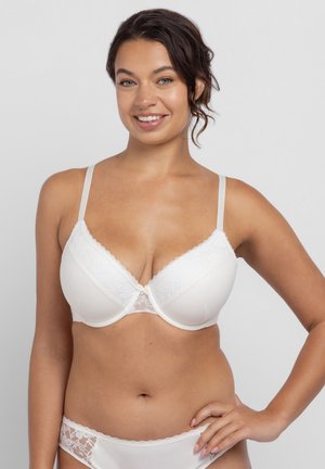 Uśmiechnięta kobieta w białym koronkowym biustonoszu i dopasowanych koronkowych majtkach, stojąca z ręką na biodrze na tle jednolitego szarego tła.