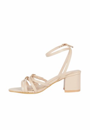 Sandal beige con tacco a blocco, caratterizzato da più cinturini incrociati e una cinghia alla caviglia. Texture liscia con dettagli dorati.