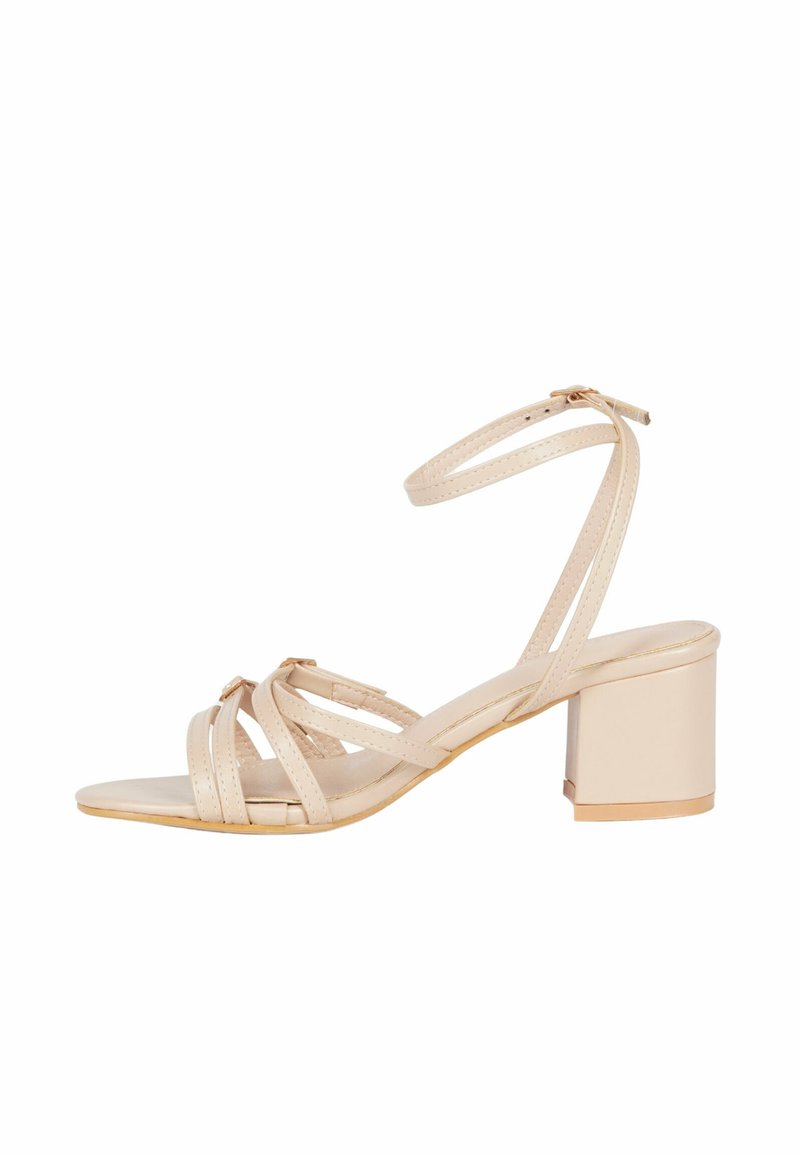Sandal beige con tacco a blocco, caratterizzato da più cinturini incrociati e una cinghia alla caviglia. Texture liscia con dettagli dorati.