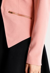 Veste ajustée rose clair avec un ourlet noir diagonal et une poche latérale ornée d'une fermeture éclair dorée. Texture lisse, manches longues.