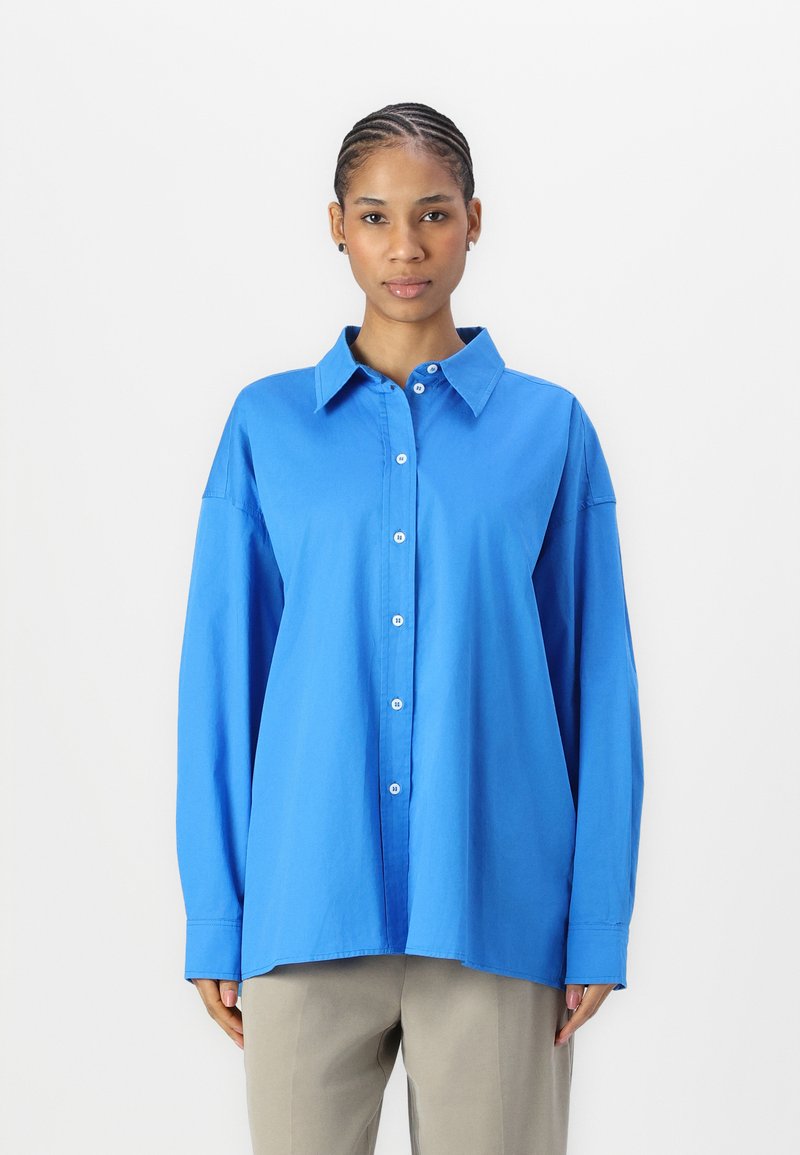 Camicia blu con colletto, maniche lunghe e vestibilità rilassata; realizzata in tessuto morbido con chiusura a bottoni visibile e polsini.