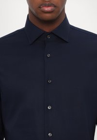 Marineblå skjorte, knappet design, lange ermer, subtil tekstur, sorte knapper, brodert "MK" logo på brystet.