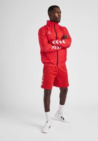Roter Sportanzug bestehend aus einer Reißverschlussjacke mit weißen Chevron-Mustern und passenden Shorts, kombiniert mit weißen Turnschuhen und Knöchelsocken.