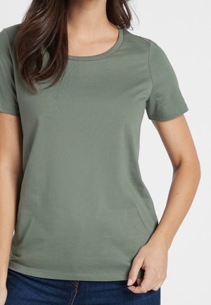Femme portant un t-shirt uni vert olive à manches courtes et un jean bleu foncé, avec une main reposant près de sa hanche.
