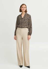 Leopardenmuster-Bluse in Braun und Schwarz, mit Kragen, kombiniert mit beigen, hoch taillierten Hosen und spitzen schwarzen Absätzen.