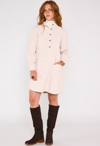 Abito in corda beige chiaro con colletto alto, chiusura frontale con bottoni e due tasche anteriori. Indossato con stivali in suede marrone al ginocchio.
