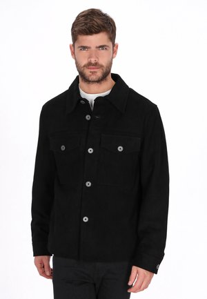 Férfi fekete, gombos kabátban, két mellzsebbel, fehér ing alatt, sima fehér háttér előtt áll.