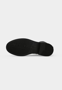 Semelle de chaussure noire avec motifs de grip texturés et nom de marque gravé sur un fond clair uni.