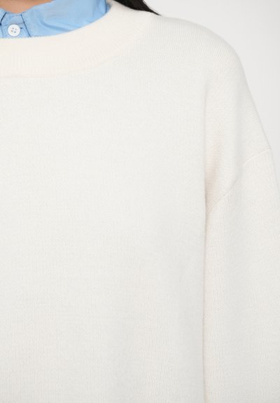 Fehér kötött pulóver kerek nyakkivágással, sima textúrával és lehajtott vállráccsal. Világoskék galléros ing fölött viselve.
