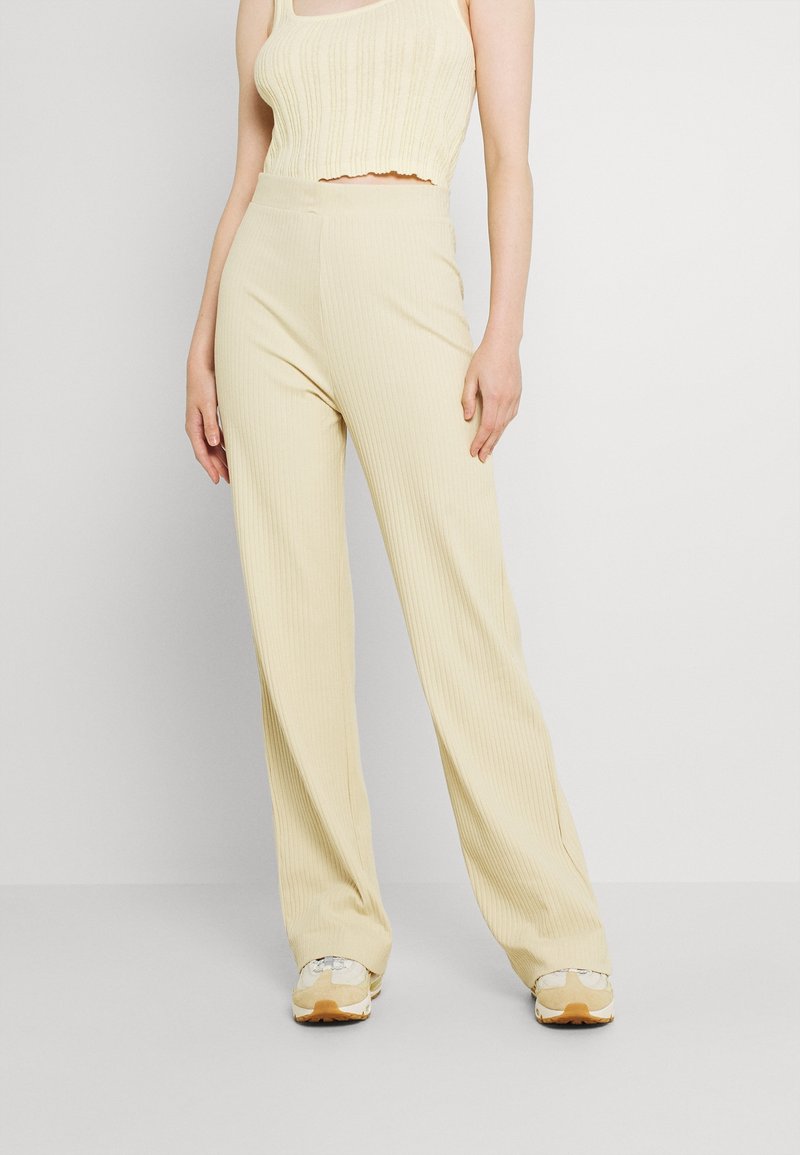 Femme portant un pantalon large côtelé jaune pâle, un débardeur court assorti et des baskets beiges, debout devant un fond blanc uni.