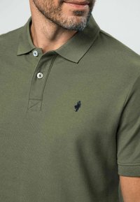 Hombre con camisa polo verde oliva con dos botones abrochados y pequeño logo negro bordado en el pecho, mostrando barba y cuello.