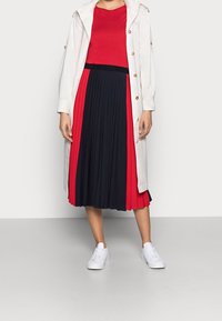 Witte trenchcoat met gouden knopen, rode top en geplooide zwart-rode rok. G gedragen met witte sneakers. Gladde textuur, gelaagd ontwerp.