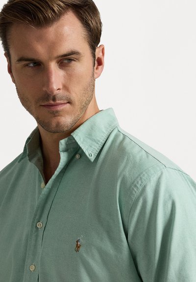 Gai zaļš pogājamais krekls ar teksturētu audumu. Iezīmēts ar apkakli, frontālajām pogām un mazu izšūtu zirga logo uz krūts.