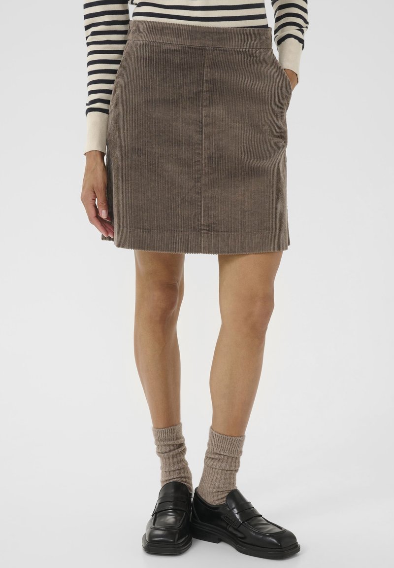 Femme portant une jupe en velours côtelé marron, un haut à manches longues rayé, des chaussettes beiges et des mocassins en cuir noir, debout avec une main dans la poche.