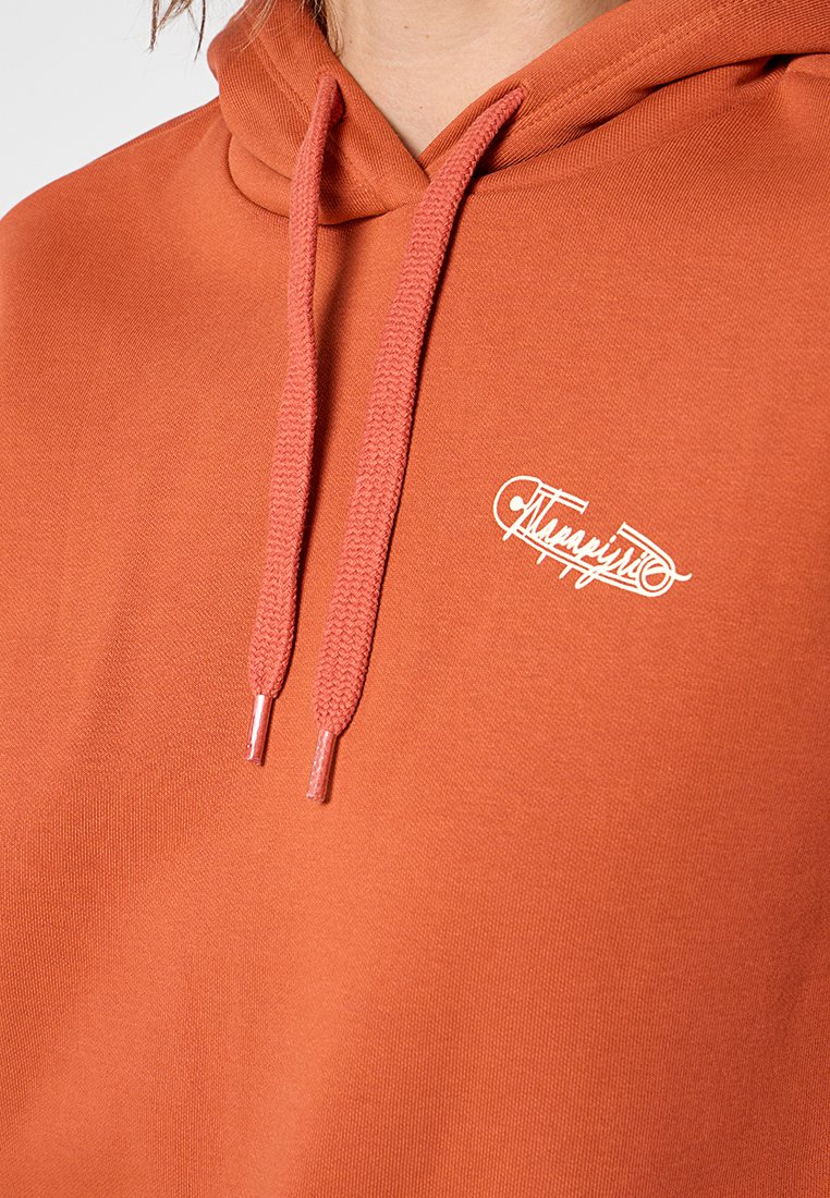 Zbliżenie na osobę w rudym sweatshirtcie z pasującymi sznurkami i małym białym logo w formie napisu na klatce piersiowej.