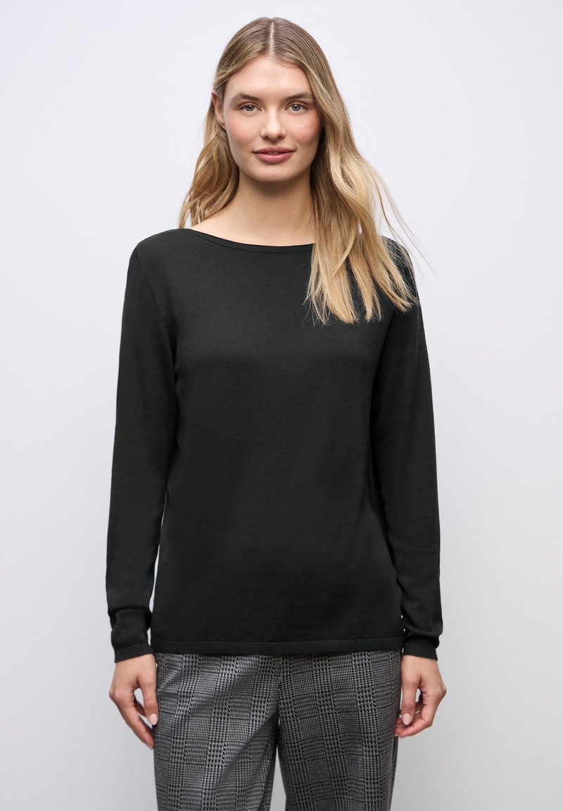 Donna con capelli biondi lunghi, indossa una maglia nera a maniche lunghe e pantaloni grigi a quadri, in piedi davanti a uno sfondo grigio chiaro uniforme.