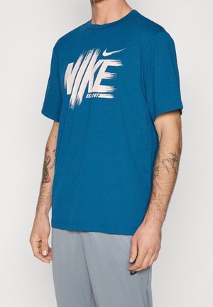 Mand iført blå Nike T-shirt med hvid penselstrøg-logo og lysegrå bukser, der viser tatoveringer på begge underarme.