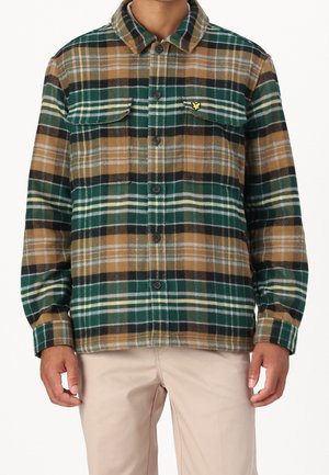 Mann trägt ein kariertes Hemd mit Knöpfen in Grün, Braun, Schwarz und Weiß mit Brusttaschen und beigefarbene Hose, gezeigt von Hals bis Hüfte.